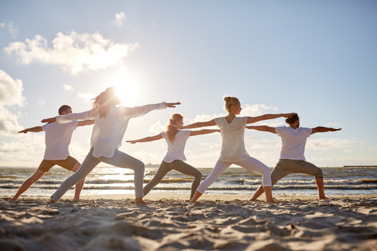 Eine Gruppe macht eine Yogaübung am Strand, alle hell gekleidet, im Sand stehend, im Hintergrund das Meer mit aufgehender Sonne